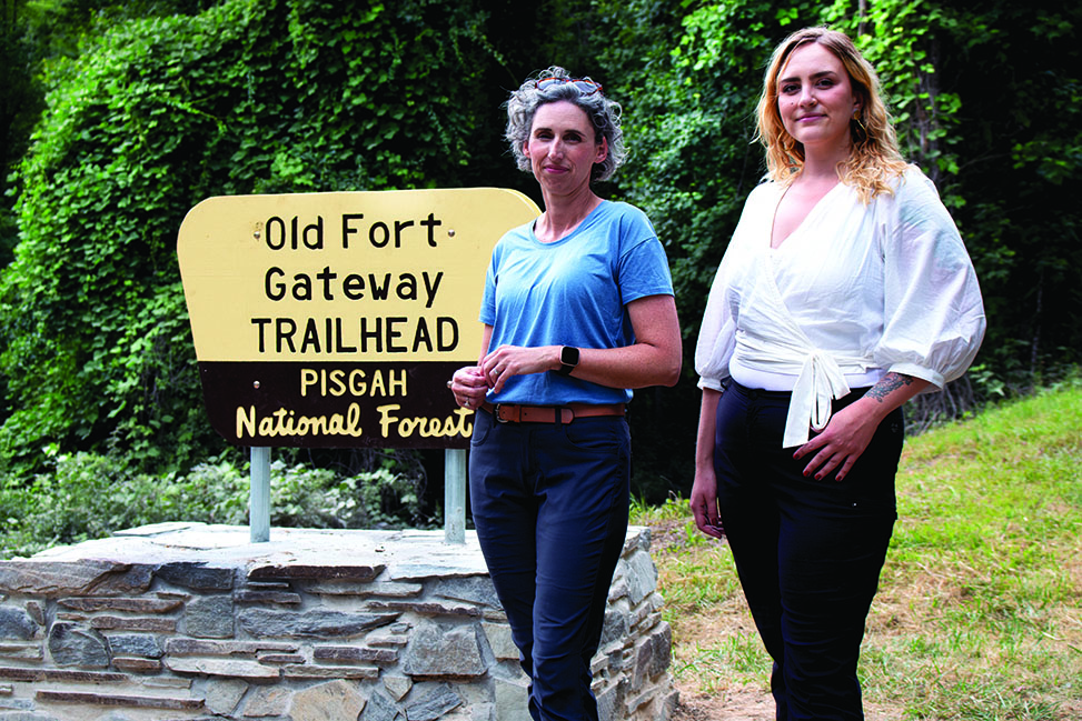 Jen Gates-Foster stands next to a black and yellow sign that reads "Old Fort Gateway Trailhead. Pisgah National Forest" and a woman named Kayla.