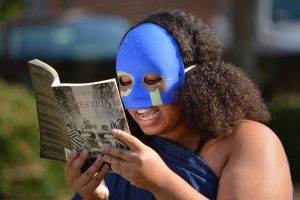 Taylor Holman in a blue mask reading from a copy of Aechylus' Oresteia.